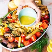 A hand pouring the salad dressing over the veggies and chicken that are grilled and layered over the greens to make the grilled chicken salad.