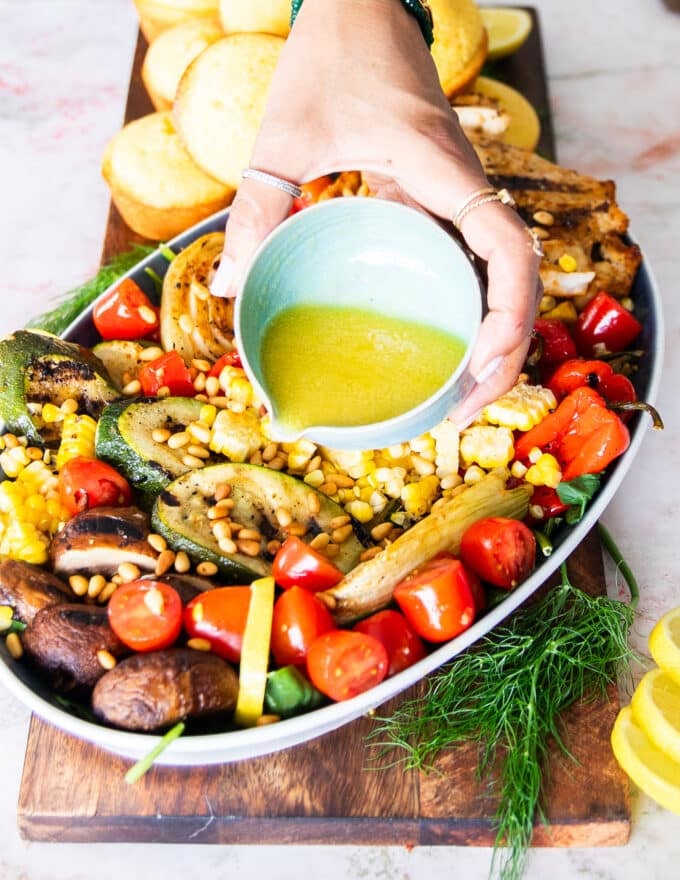 A hand pouring the salad dressing over the veggies and chicken that are grilled and layered over the greens to make the grilled chicken salad.