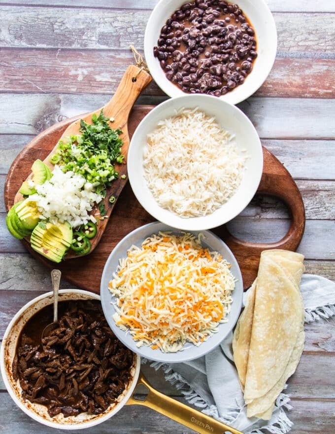 Ingredients needed to make birria burritos in a large board including birria meat, some cheese, tortillas, beans, rice, onions, cilantro and consommé 