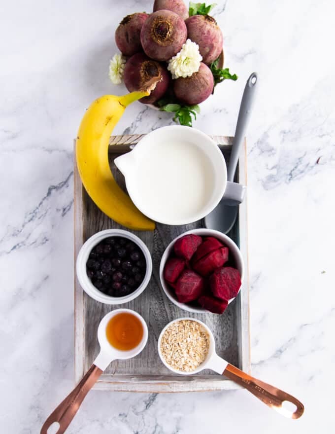 Ingredients for a beet smoothies in a tray showing fresh beets cut up, a banana some milk, honey, and frozen blueberries